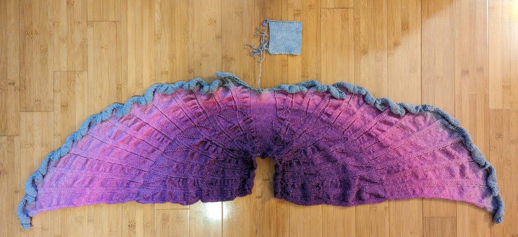 A shawl with a feather motif.  The cowl is a deep purple that fades out to fuchsia before it then blends to silver for the feather tips. There is some silver thread in the yarn giving little silver sparkles. It is fresh off the needles and therefore needs to be blocked.  It is laying on a wood floor.