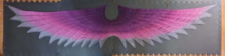 A shawl with a feather motif. The cowl is a deep purple that fades out to fuchsia before it then blends to silver for the feather tips. There is some silver thread in the yarn giving little silver sparkles. It is pinned to a mat.