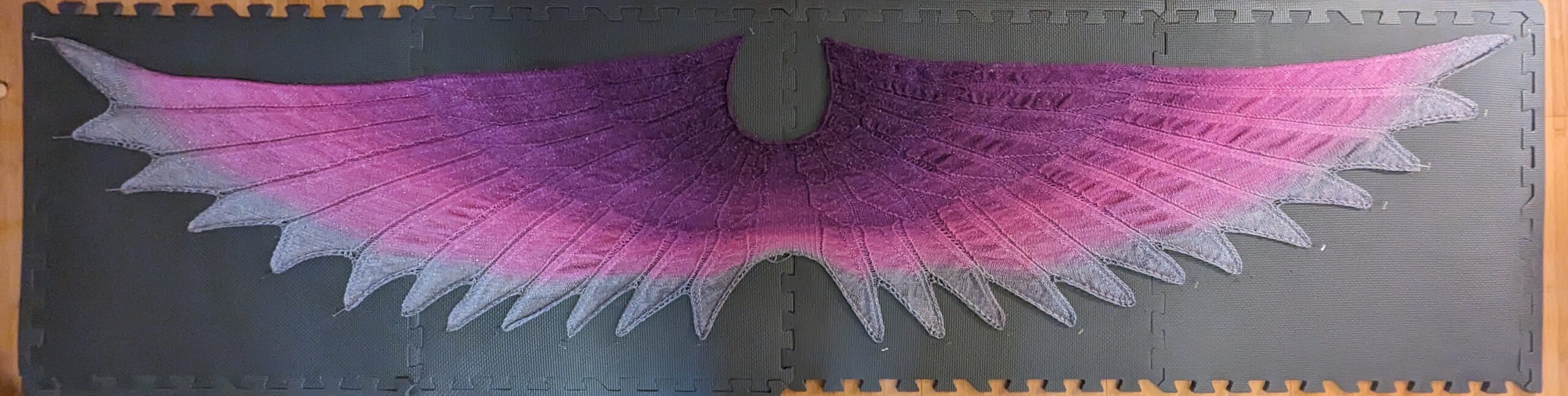 A shawl with a feather motif. The cowl is a deep purple that fades out to fuchsia before it then blends to silver for the feather tips. There is some silver thread in the yarn giving little silver sparkles. It is pinned to a mat.