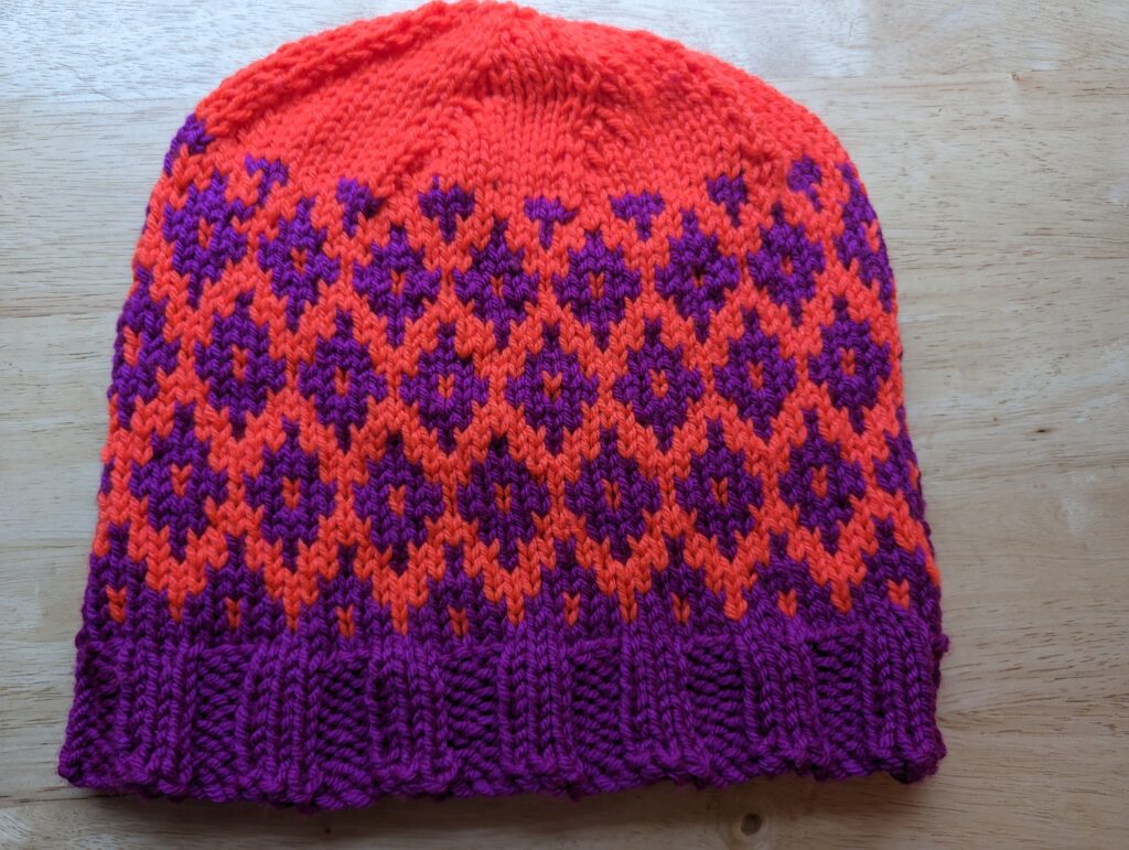 A colorwork hat in violet and neon orange with a diamond patterns.  The rim is violet and the crown is orange.