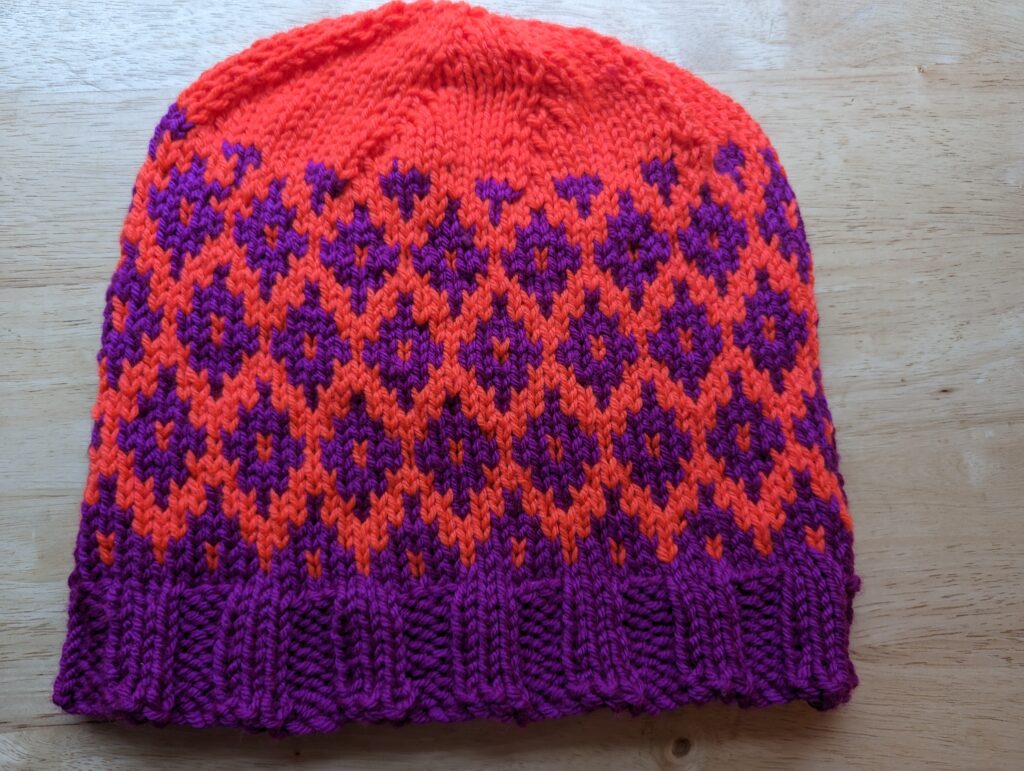 A colorwork hat in violet and neon orange with a diamond patterns. The rim is violet and the crown is orange.