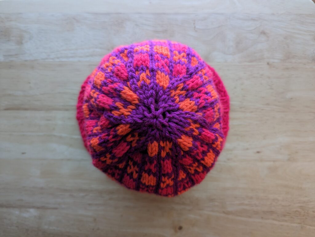 top down view of a pink brimmed and pink and orange alternating checkerboard block pattern with violet bordering. Violet yarn completes the crown.