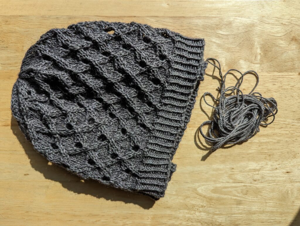 A folded slouchey berey with cables around syncopated holes atop a twist knit band next to some leftover purplish grey yarn